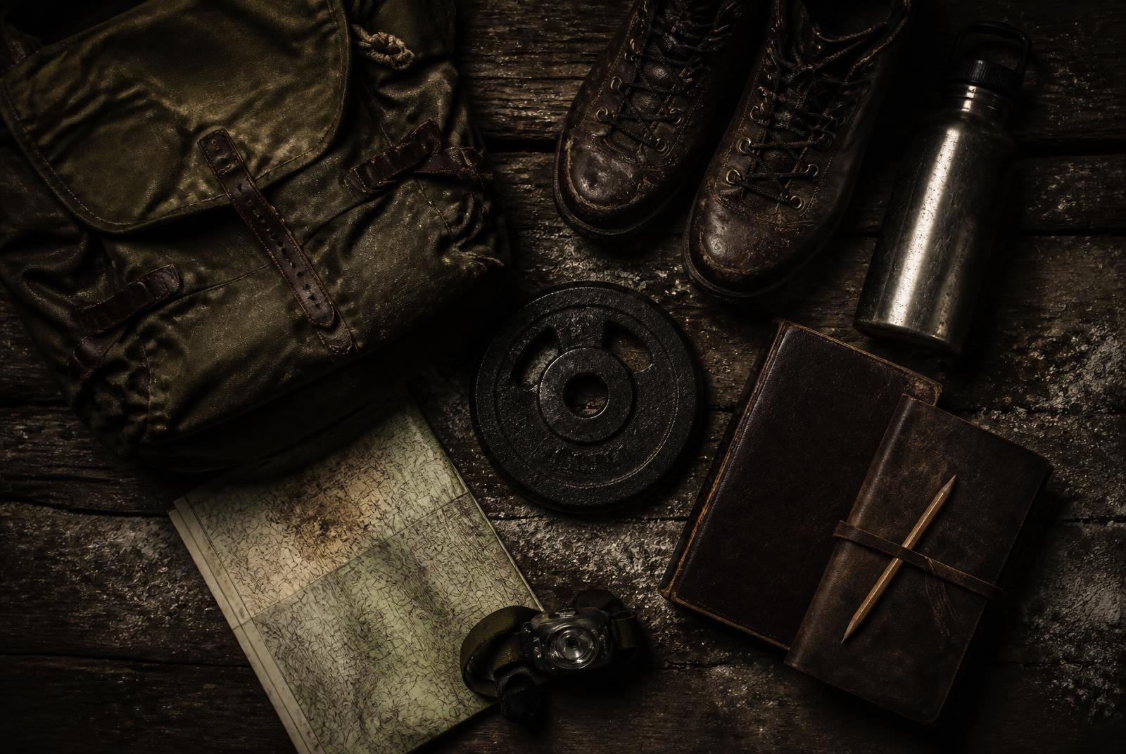 Overhead view of ruck, worn boots, water bottle, weight plate, Bible, journal and topographic map laid out on weathered wood